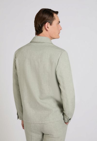 Mężczyzna o krótkich brązowych włosach nosi jasnooliwkową marynarkę i spodnie, stoi bokiem z rękami w kieszeniach na jednolitym tle.