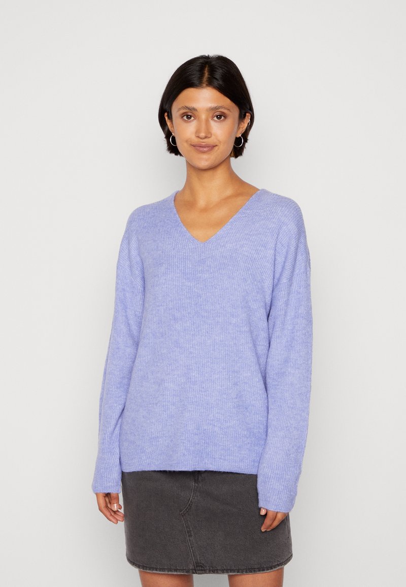 Pull col en V violet clair avec des manches longues, texture côtelée et coupe décontractée, porté sur une jupe en denim foncé.