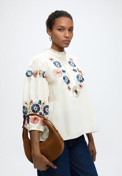 Femme portant un chemisier blanc avec une broderie florale multicolore, tenant un sac à main marron en forme de croissant, debout devant un fond uni.