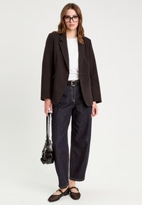 Blazer noir sur un haut blanc, associé à un denim sombre, une ceinture noire et des chaussures en mesh noires. Les accessoires comprennent un petit sac noir.