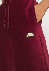 Pantalon en velours côtelé bordeaux avec une finition texturée, présentant une taille à cordon de serrage et un patch de logo rehaussé d'or sur la cuisse.
