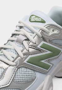 Αθλητικό παπούτσι New Balance 9060 σε λευκό και ασημί με λευκά κορδόνια και πράσινες λεπτομέρειες λογότυπου, με κοντινή όψη από το πλάι και από πάνω.