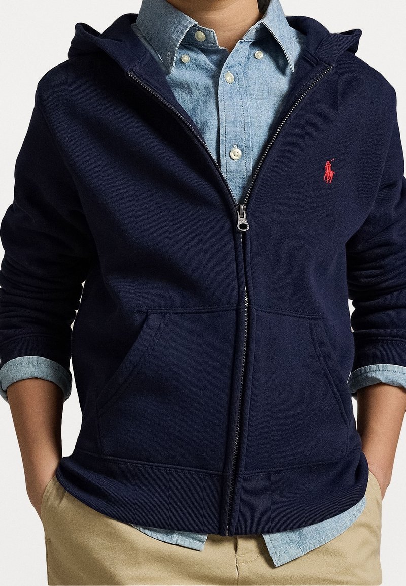 Marinblå zip-up hoodie i mjukt material, med framsida dragkedja, två fickor och en röd logga på bröstet. Lagd över en denimknäppt skjorta.