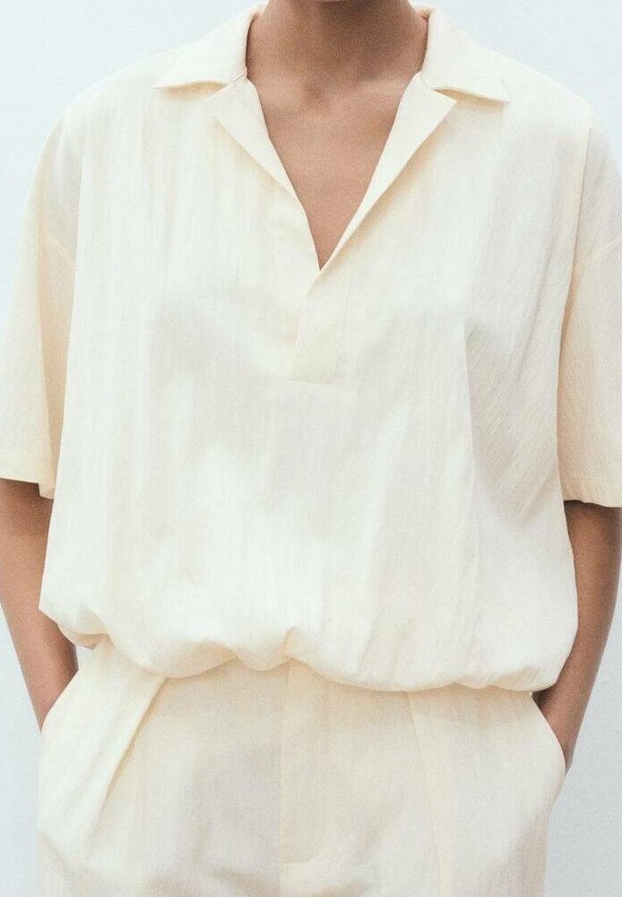 Personne portant une chemise ample crème à manches courtes avec col et col en V, rentrée dans un pantalon taille haute assorti, les mains dans les poches.