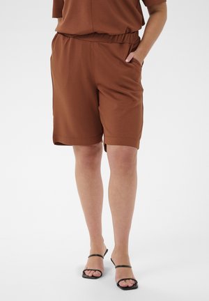 Osoba ubrana w brązowe krótkie spodenki do kolan i czarne sandały na obcasie z odkrytymi palcami, stojąca z jedną ręką w kieszeni na jednolitym tle.