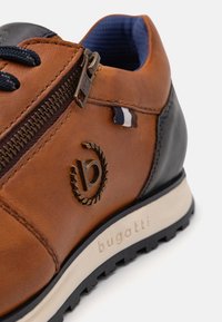 Baskets en cuir marron et noir avec fermeture éclair latérale, lacets foncés, doublure intérieure bleue, logo Bugatti, et semelle en caoutchouc blanche avec motif de crampons.