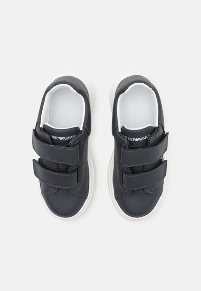 Mörkblå sneakers med två kardborreband, vita gummisålar, texturerade detaljer på hälen och en slät ytfinish.