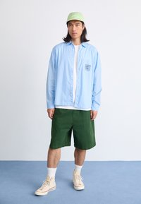 Camisa de botón a rayas azul claro con "OBEY" en el lado izquierdo del pecho, combinada con pantalones cortos verdes y zapatillas de caña alta beige. Gorra verde claro.