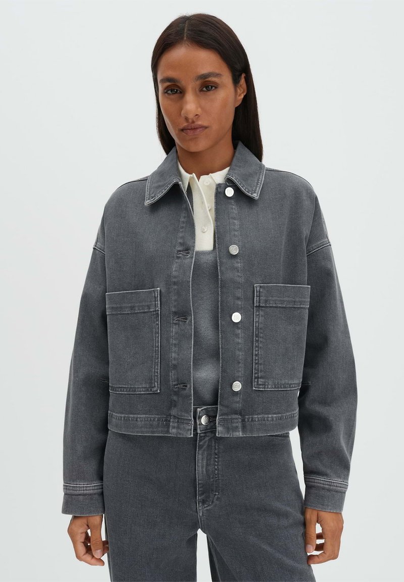 Vrouw draagt een cropped grijze spijkerjas met grote voorzakken en bijpassende hoge taille grijze spijkerbroek, witte blouse eronder.