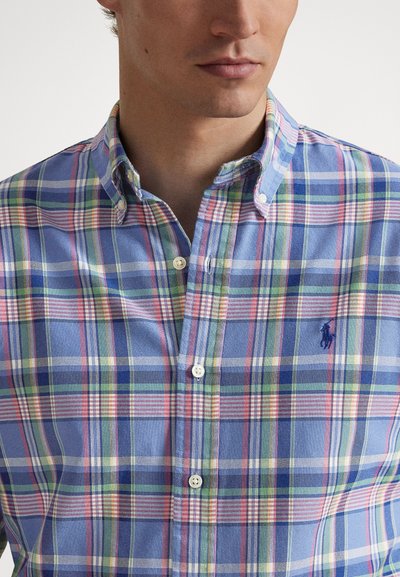 Skjorta med knappar i blått och pastellrutigt mönster; har krage och logotyp. Materialet verkar lätt och andningsbart.