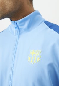 Casaco desportivo azul claro com colarinho alto, apresentando o logo amarelo do FC Barcelona e detalhes em azul escuro nos ombros. Tecido liso e leve.
