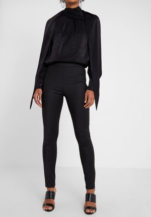 Vrouw die zwarte, hooggetailleerde, smalle broek draagt, een zwart, lang mouwtop met brede mouwen, zwarte open-toe hakken en opvallende oorbellen.