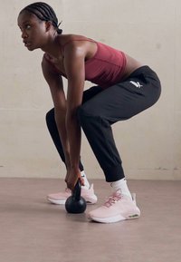Crni kettlebell drži osoba u tamnocrvenoj sportskoj majici i crnim hlačama, koja nosi ružičaste tenisice, bijele čarape i pletenu kosu.