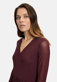 Burgunderfarbenes Kleid mit V-Ausschnitt, langen transparenten Ärmeln und einem plissierten Rock. Der Stoff hat eine glatte Textur und ein tailliertes Oberteil.