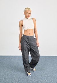 Szürke cargo nadrág oldalsó zsebekkel, egy bordázott krémszínű crop top-pal párosítva. A modellen tengerészkék és sárga sportcipők vannak, és egy kék szőnyegen áll.