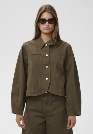 Femme portant une veste boutonnée vert olive foncé et un pantalon assorti avec des lunettes de soleil ovales noires sur fond uni.