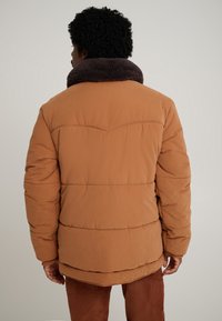 Casaco acolchoado em cor ferrugem com gola em pelo falso castanho escuro texturizado. Apresenta costuras horizontais e punhos canelados.