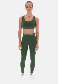 Alyvuogių žalios spalvos sportinė liemenėlė ir tamprių komplektas su kontrastinga juosmens juosta. Tamprės prigludusios, medžiaga atrodo tampri. Baltos sportinės bateliai užbaigia įvaizdį.