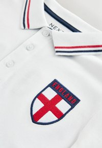 Polo blanc avec col rayé rouge et marine et logo écusson d’Angleterre brodé sur la poitrine.