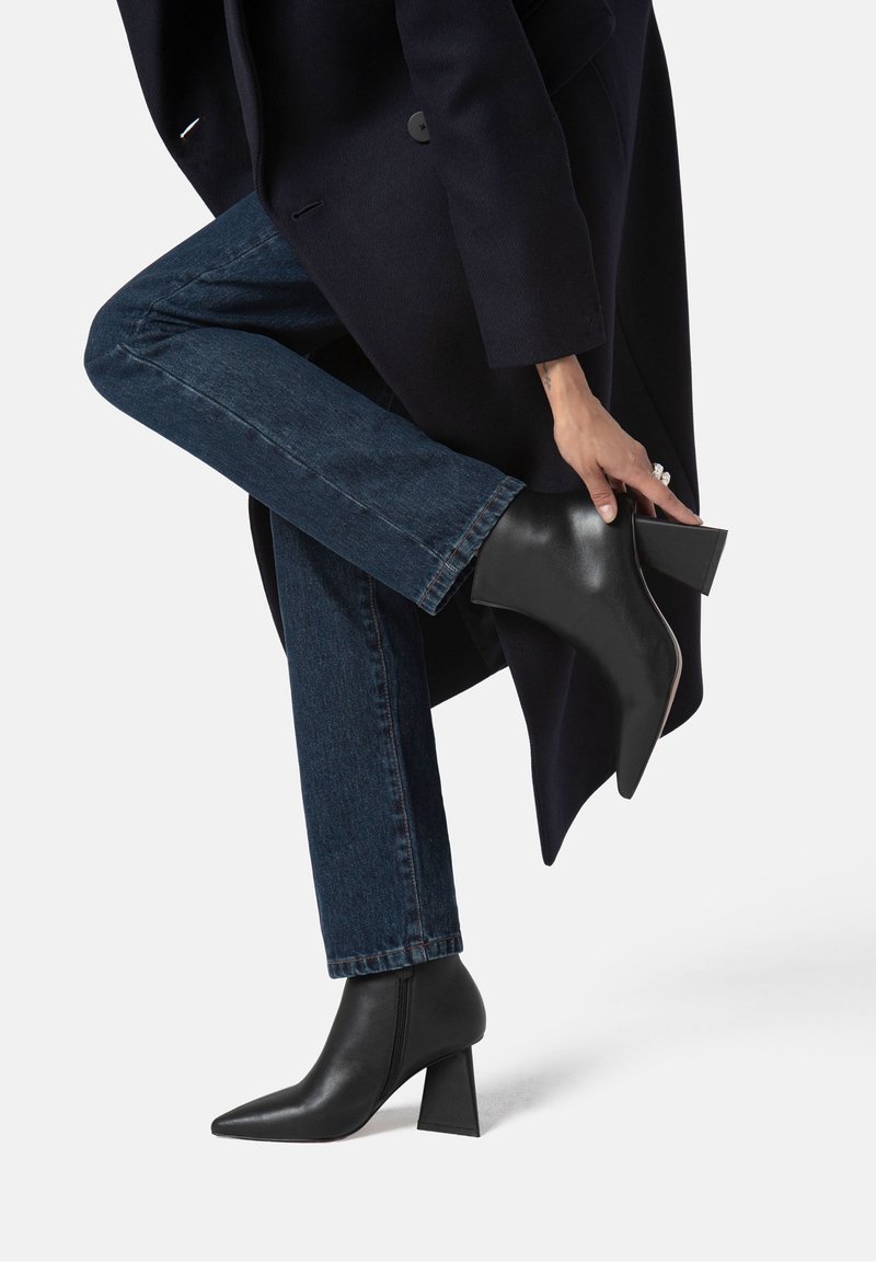 Personne portant un jean foncé et des bottines noires à talons, levant une jambe tout en ajustant la botte, vêtue d'un manteau sombre.