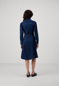 Mörk denimklänning med en åtsittande överdel, puffärmar och en utställd kjol. Har en bälte i midjan och knäppning vid ärmsluten.