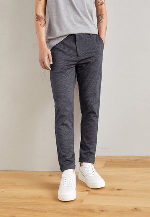 Pantalon gris chiné avec une coupe slim, taille élastique et poches latérales, associé à des baskets blanches sur un sol en bois.