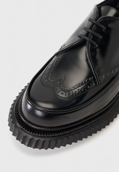 Chaussure habillée en cuir noir avec détails à bout golf et semelle en caoutchouc épaisse et nervurée, vue de face sur un fond pâle.