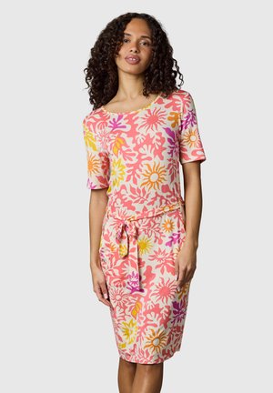 Kvinde med krøllet hår iført en knælængde, kortærmet kjole med pink, orange, gul og lilla sol- og bladmønstre, bundet ved taljen.
