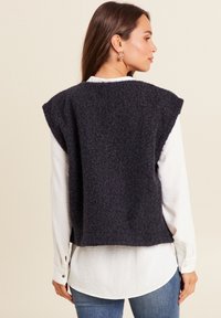 Dunkelgraue Wollweste mit strukturiertem Finish, breitem Schulterndesign und kurzem Schnitt, getragen über einem weißen Hemd und kombiniert mit blauen Jeans.