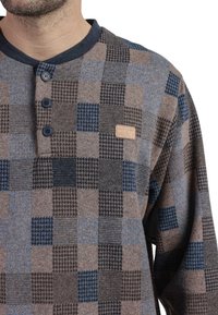 Camisa de botones, de manga larga, en un patrón de cuadros marrones, azules y grises. Presenta un cuello oscuro y un pequeño parche de logo de cuero.