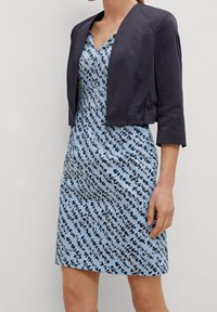Marinblå kort jacka över en blå klänning med ett spritt svart- och vitt mönster. Klänningen har V-ringning och en figurnära design.