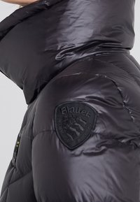 Gros plan sur l'épaule et le col haut d'une doudoune noire avec un patch de la marque Blauer sur la manche.