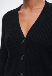 Gros plan d'une personne portant un cardigan noir côtelé boutonné avec un décolleté en V profond.