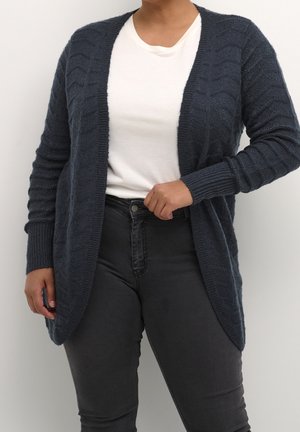 Person wearing a dark blue textured knit cardigan over a white t-shirt and high-waisted dark gray jeans, standing against a plain background.