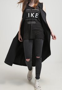 Svart ärmlös topp med "LIKE" grafiskt tryck, lager på lager över en lång svart kappa; kombineras med slitna svarta jeans och vita sneakers.