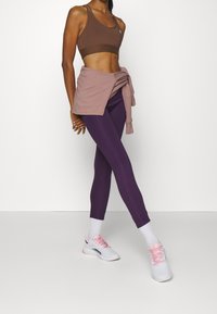 Hnědá sportovní podprsenka s širokým pásem, spárovaná s vysokými fialovými legínami. Světle růžový svetr uvázaný v pase a bílé sportovní boty.