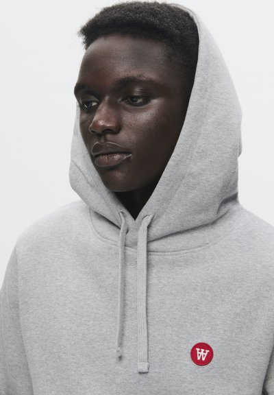 Grauer Kapuzenpullover mit einer vorderen Kängurutasche, der ein kleines rotes kreisförmiges Logo zeigt. Der Stoff ist weich und strukturiert.