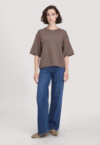 Maglione marrone a trama con maniche corte, abbinato a jeans in denim blu a gamba larga e scarpe piatte beige, in piedi davanti a uno sfondo chiaro.