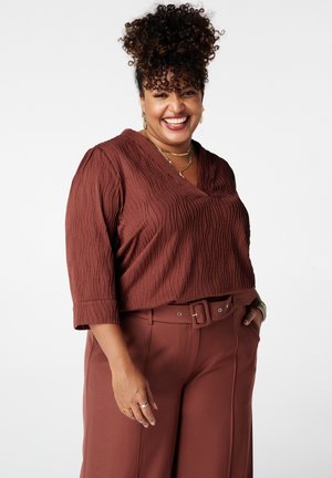 Lachende vrouw met krullend haar, gekleed in een getextureerde roestkleurige blouse en bijpassende broek met ceintuur, voorzien van gouden sieraden en ringen.