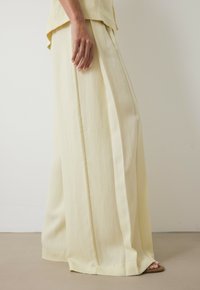 Pantalons à jambes larges en tissu jaune doux, présentant une texture lisse, des plis et un drapé confortable. Associés à un haut assorti.