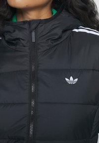 Chaqueta de plumas negra con cuello alto, cremallera frontal y el logo de Adidas en blanco en el lado izquierdo del pecho. Presenta costuras horizontales y una textura suave.