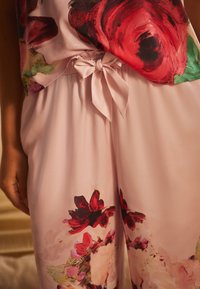 Blumenbluse mit lebhaften roten und grünen Mustern, kombiniert mit hellrosa Hosen, die ähnliche Blumenakzente und eine Schleife am Taillenbund haben.
