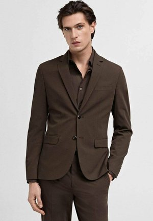 Completo marrone su misura con giacca aderente, rever a punte e due tasche frontali. Abbinato a una camicia dress marrone coordinata. Texture liscia.