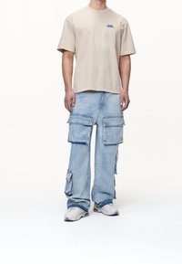 Beige bomullströja med korta ärmar och blå logotyp, ihop med ljusblå cargobyxor med stora fickor och slitningar. Vita sneakers.