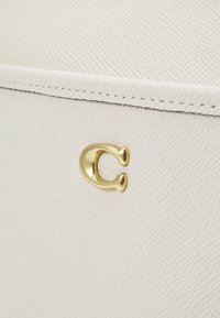 Primo piano di una borsa in pelle bianca, con una finitura leggermente testurizzata e un accento hardware a forma di C dorata sul lato anteriore.