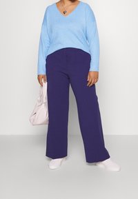 Lichtblauwe oversized trui met V-hals, gecombineerd met wijde paarse broek. Een zachtroze handtas met knoopdetail wordt vastgehouden. Witte sneakers.