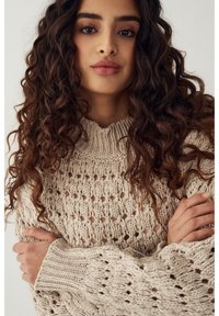 Noor naine pikkade lokkis tumedate juustega, seljas beež kootud kampsun, käed rüpes, neutraalne väljendus, kerge ühevärviline taust.