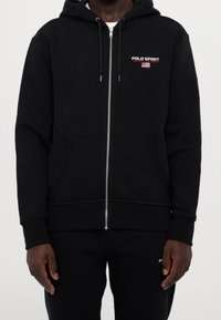 Černá mikina na zip z bavlněné směsi. Má kapuci, dvě přední kapsy a na hrudi nápis "POLO SPORT" s doprovodnou grafikou.