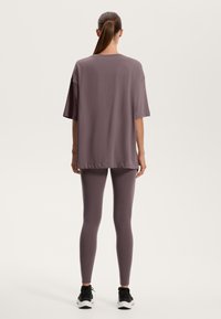 Přehozené fialové tričko s krátkým rukávem a zakulaceným lemem, spárované s přiléhavými fialovými legínami a černými sportovními botami.