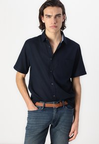 Chemise à manches courtes bleu marine foncé avec un col boutonné, une poche poitrine unique et une texture douce, associée à un jean bleu et une ceinture brune.